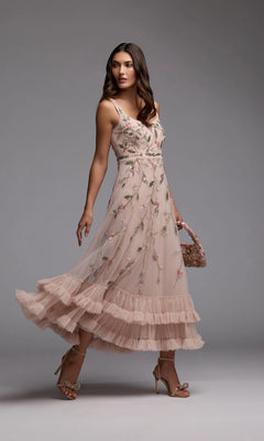 A woman stands against a plain gray background wearing the Mac Duggal 9402 long formal dress—a light pink, sleeveless party gown adorned with delicate floral beaded embroidery and featuring a ruffled hem. She completes the elegant ensemble with a matching handbag.