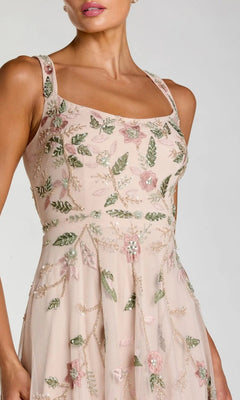 A woman is shown from shoulders to knees against a plain background, wearing the Mac Duggal 9402 long formal dress—a sleeveless, light pink party gown adorned with green and pink floral beaded embroidery.