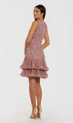 A woman wearing the Mac Duggal 9403 Short Party Dress—a sleeveless, knee-length pink fit and flare design featuring floral embroidery and tiered ruffles—faces slightly away while pairing it with gold high-heeled sandals against a plain white background.