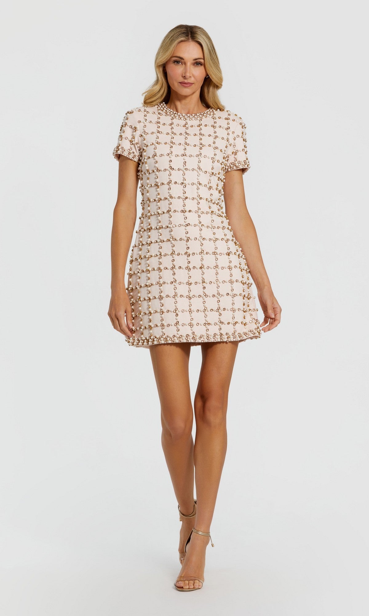 A woman wearing the Mac Duggal 94036 Short Party Dress in blush—a short-sleeved mini dress featuring a geometric pattern with subtle pearl and rhinestone embellishments—stands against a plain white background in sandals.