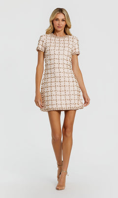A woman wearing the Mac Duggal 94036 Short Party Dress in blush—a short-sleeved mini dress featuring a geometric pattern with subtle pearl and rhinestone embellishments—stands against a plain white background in sandals.