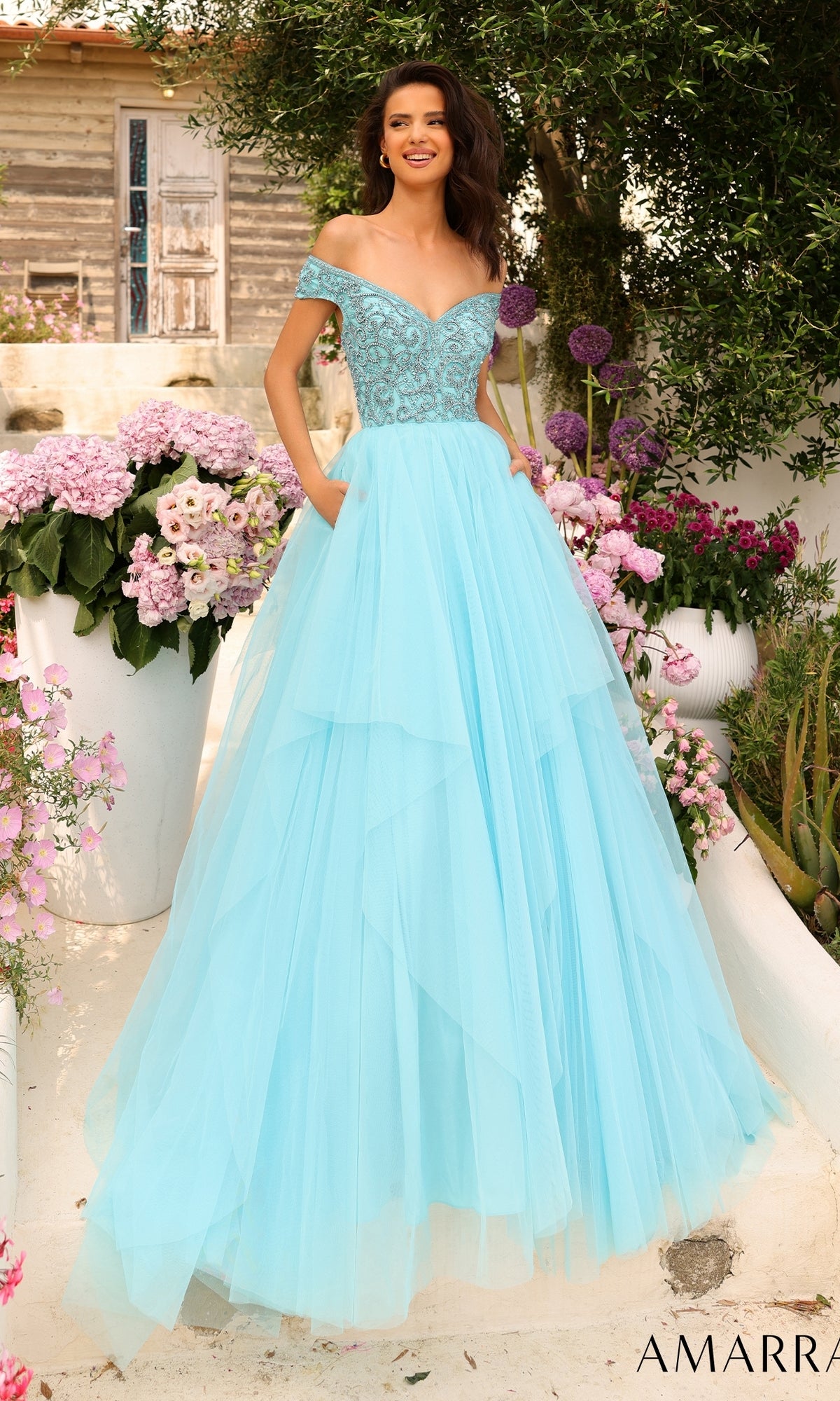 A woman smiles outdoors among pink and purple flowers, wearing the Beaded-Bodice Long A-Line Prom Dress: Amarra 94038 in light blue with an off-the-shoulder design.