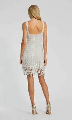 A woman faces away, wearing the White Pearl Fringe Cocktail Dress by Mac Duggal 94049—sleeveless, white, with a fringed hemline—paired with metallic high heels against a plain background.