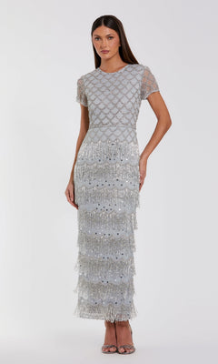 A woman stands against a plain white background wearing the Mac Duggal 94077 Fringed Short Sleeve Party Dress—a silver, floor-length gown with beaded geometric patterns and tiered fringe.