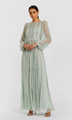 A woman wearing the Mac Duggal 94081 long sleeve formal dress in light green with sheer, beaded sleeves stands with hands clasped, looking at the camera.