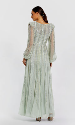 A woman stands facing away, wearing the Mac Duggal 94081—a modest, floor-length pale green formal dress with long sheer sleeves and vertical sequin details.
