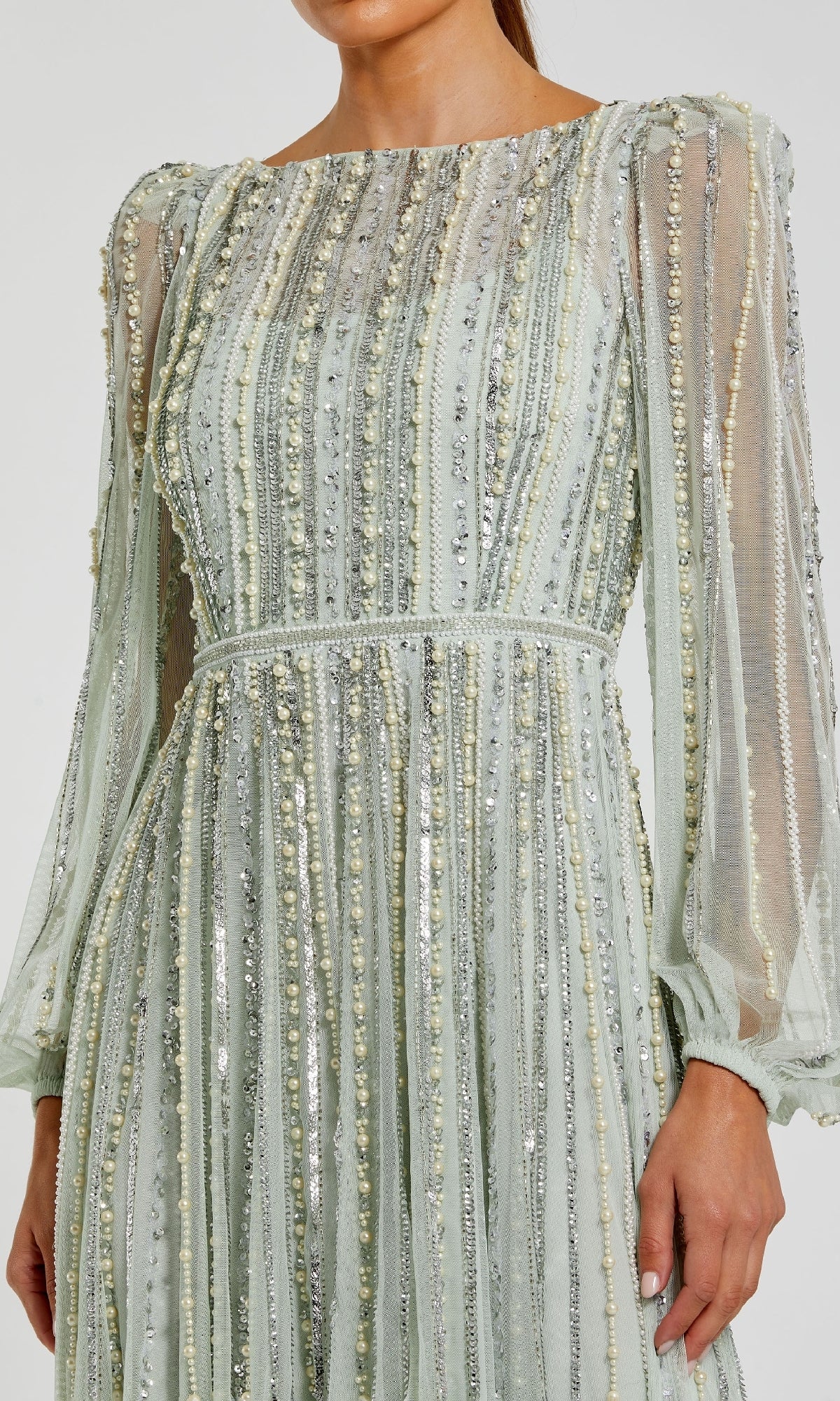 A woman wears the Mac Duggal 94081, a long sleeve formal dress in light green with sheer fabric and vertical metallic sequin and bead embellishments.