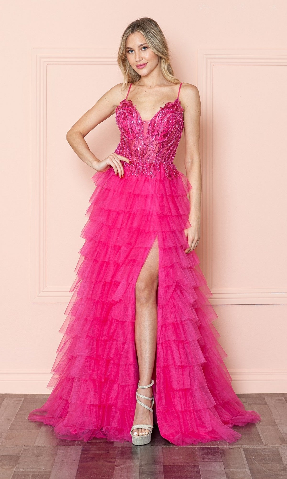 A woman models the Open-Back Long Ruffled Prom Dress: Poly USA 9408, featuring a bright pink hue, spaghetti straps, beaded v-neck bodice, and high slit, standing by a light pink wall and wooden floor.