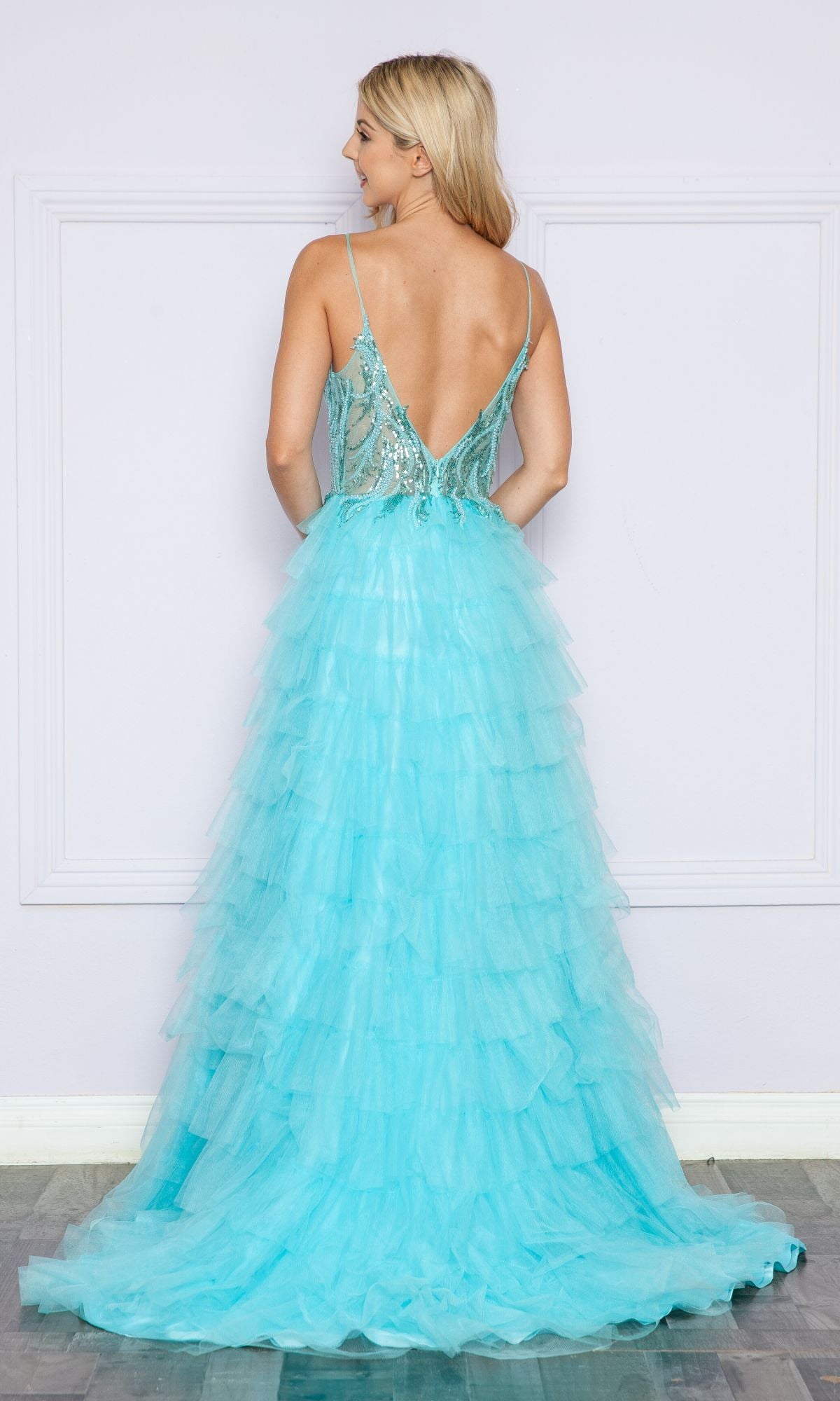 A woman faces away from the camera in the Poly USA 9408 Open-Back Long Ruffled Prom Dress, a light blue tulle gown with a deep V-back, spaghetti straps, standing by a pale wall on a wooden floor.
