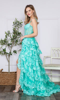 A woman stands indoors by a potted plant and white bench, wearing the Poly USA 9410 Sheer-Corset Long Ruffled Prom Dress in turquoise with a sleeveless design and high-low hem.