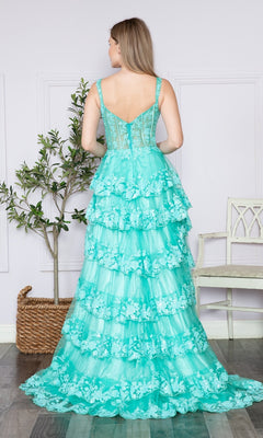 A woman in the Sheer-Corset Long Ruffled Prom Dress (Poly USA 9410), a turquoise tiered lace gown with a corset bodice, stands with her back to the camera in a bright room featuring light walls, a white chair, and a potted plant.