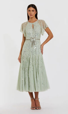 A woman stands in sandals against a plain background, wearing the Mac Duggal 94128 Tea-Length Party Dress in seafoam—a star sequin dress featuring sheer short sleeves, a keyhole neckline, and a beaded bow belt.
