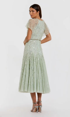 A woman stands against a plain white background, looking over her shoulder in a tea-length party dress reminiscent of the Mac Duggal 94128, featuring light green fabric with star sequins, short flutter sleeves, and paired with silver high heels.