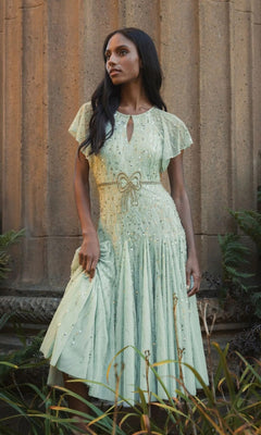 A woman in a light green, short-sleeved Tea-Length Party Dress: Mac Duggal 94128 stands outdoors near a stone column, looking to the side.