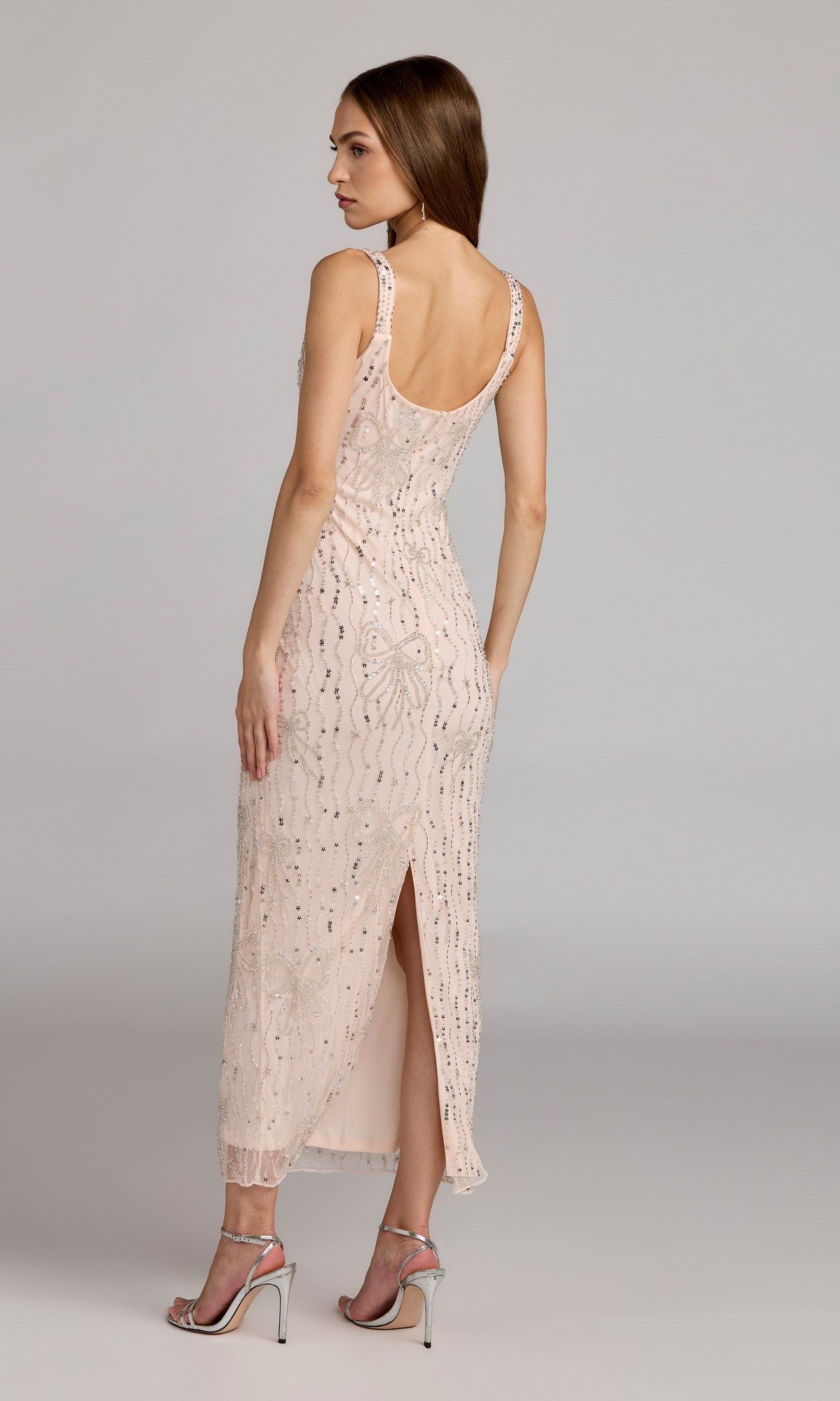 A woman faces away, wearing the Mac Duggal 94129 long formal sleeveless beaded mesh ankle-length dress with a back slit and silver high-heeled sandals against a plain background.