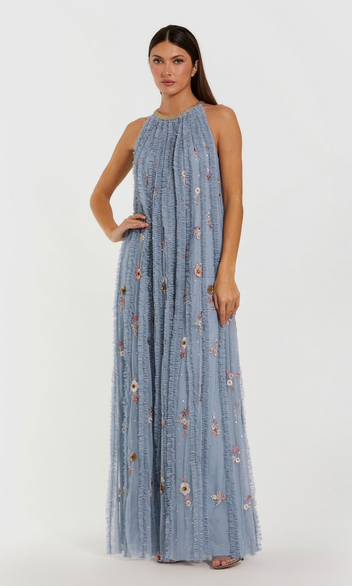 A woman wearing the Mac Duggal 9413 long formal dress in Blue Multi, featuring a sleeveless design, floor-length silhouette, ruffle details, and embroidered floral patterns, stands against a plain white background.