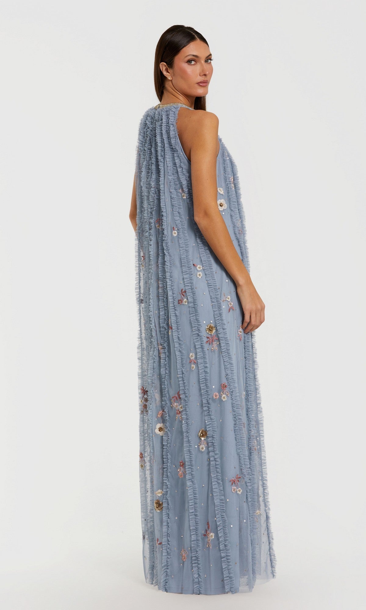 A woman wears the Mac Duggal 9413 long formal dress—a sleeveless, floor-length blue gown adorned with intricate floral embroidery—while standing and gazing over her shoulder against a plain white background.