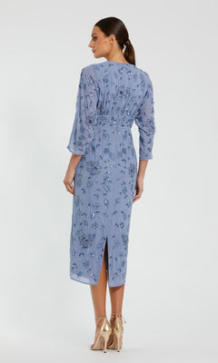 A woman stands with her back to the camera, wearing the Mac Duggal 94141 Midi Party Dress—a light blue, long-sleeve dress featuring intricate embroidery and modest cocktail styling—paired with gold high-heeled shoes against a plain white background.