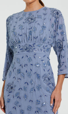 A person wearing the Mac Duggal 94141 Midi Party Dress, which features a light blue hue, intricate floral embroidery with sequin accents, three-quarter sleeves, a fitted waist, and flowy fabric. Only the upper body is visible.