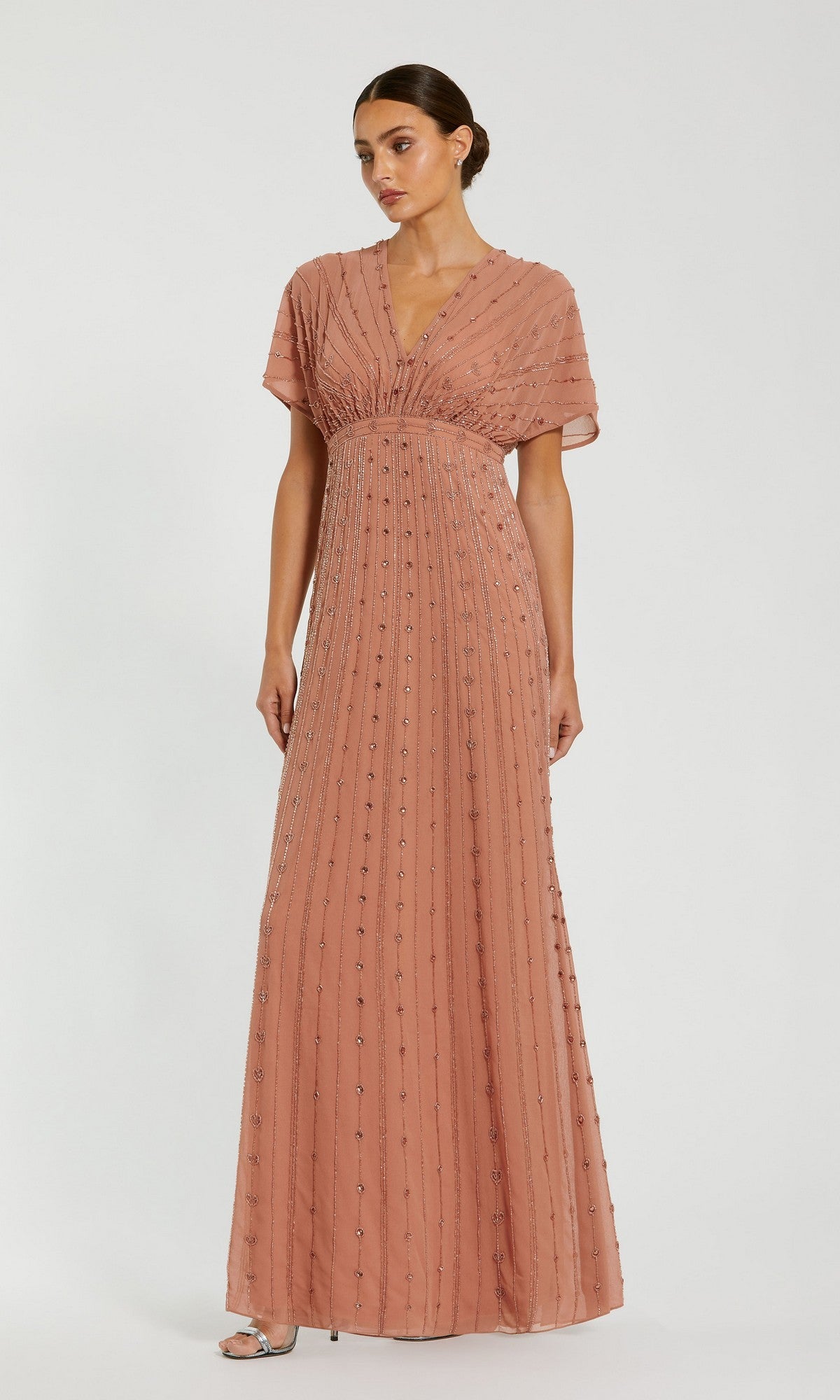 A woman stands wearing the Long Formal Dress: Mac Duggal 94149, a floor-length, cinnamon-colored embellished gown with short sleeves, a v-neckline, and beaded vertical stripes; her hair is styled in a low bun and she pairs the look with open-toed heels.