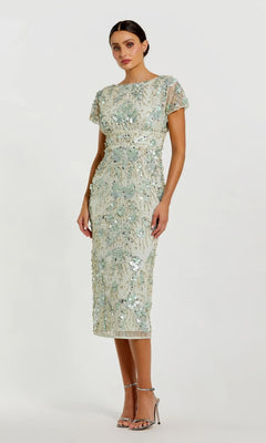 A woman wears the Mac Duggal 94150 Midi Wedding Guest Dress in sage with sparkling embellishments and silver high-heeled sandals, standing against a plain background.