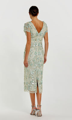 A woman faces away in a light green Mac Duggal 94150 Midi Sequin Wedding Guest Dress featuring short sleeves and a V-shaped back. Her hair is styled in a low bun, and she completes the look with elegant heels.