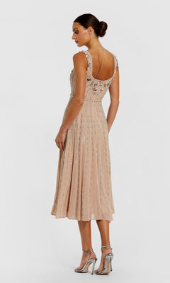 A woman wearing the Rose Midi Wedding Guest Dress: Mac Duggal 94155 in light pink with beaded embellishments and a flared skirt, paired with silver high-heeled sandals, stands against a plain background.
