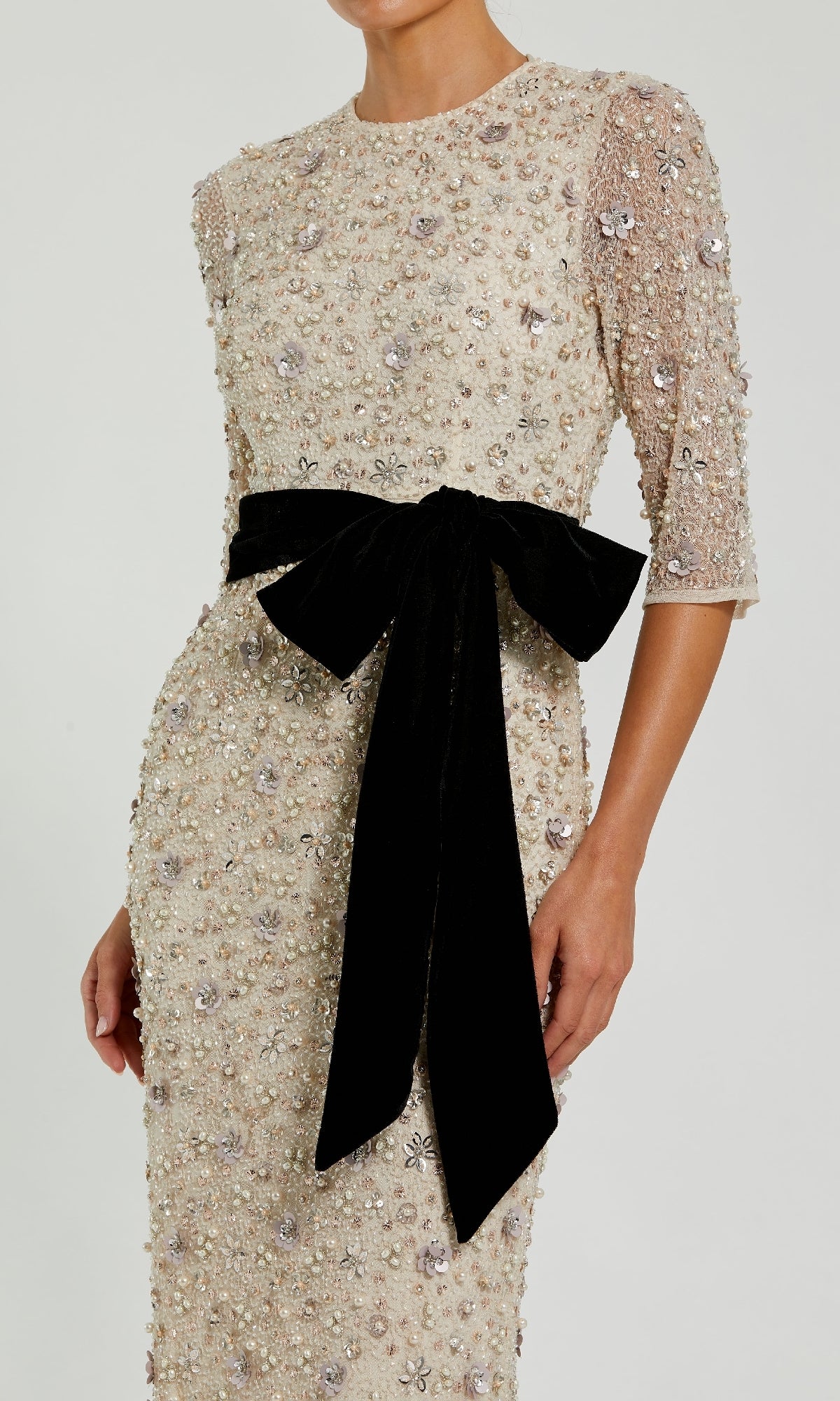 A woman wearing the Beaded Oyster White Formal Gown (Mac Duggal 94166)—a cream beaded dress with three-quarter sleeves and a black velvet waist bow—stuns at any special occasion.