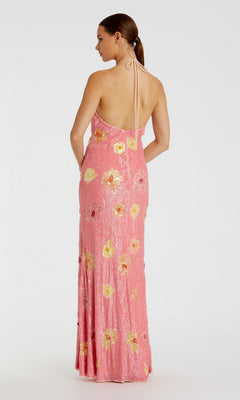 A woman is wearing the Mac Duggal 94175 Long Formal Dress, a pink halter-neck gown adorned with yellow and white floral sequins. The floor-length, backless silhouette stands out against a plain white background, radiating elegance and sophistication.