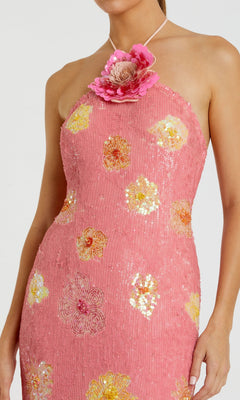 Wearing the Mac Duggal 94175 long formal dress, this person showcases a pink halter-neck design embellished with yellow and pink floral patterns, sparkling sequins, and a prominent pink flower at the neckline.
