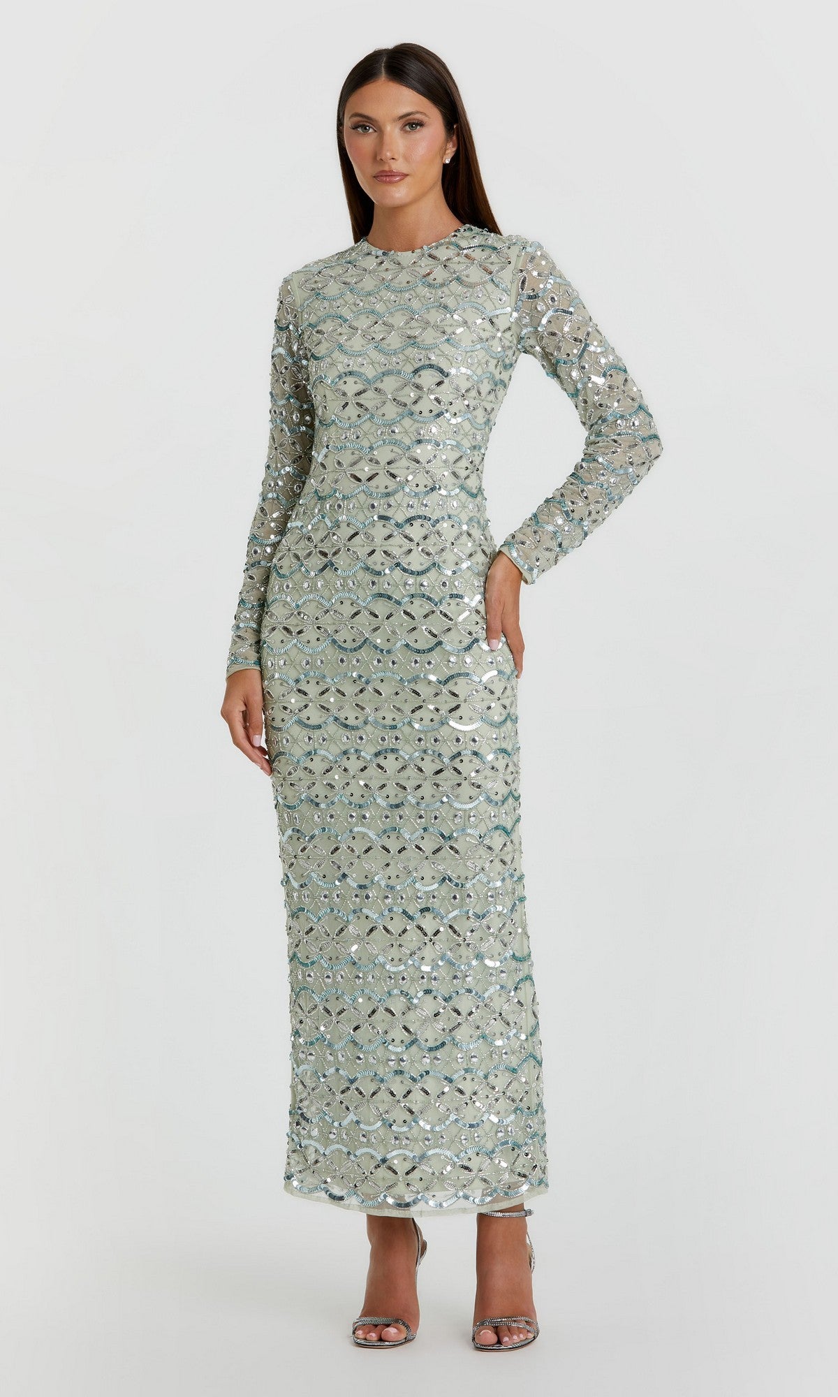 A woman stands wearing the Mac Duggal 94194 Long Formal Dress in jade, a long-sleeved, floor-length gown featuring striking silver sequins and geometric beadwork, paired with silver high-heeled sandals against a plain light background.