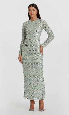 A woman stands wearing the Mac Duggal 94194 Long Formal Dress in jade, a long-sleeved, floor-length gown featuring striking silver sequins and geometric beadwork, paired with silver high-heeled sandals against a plain light background.