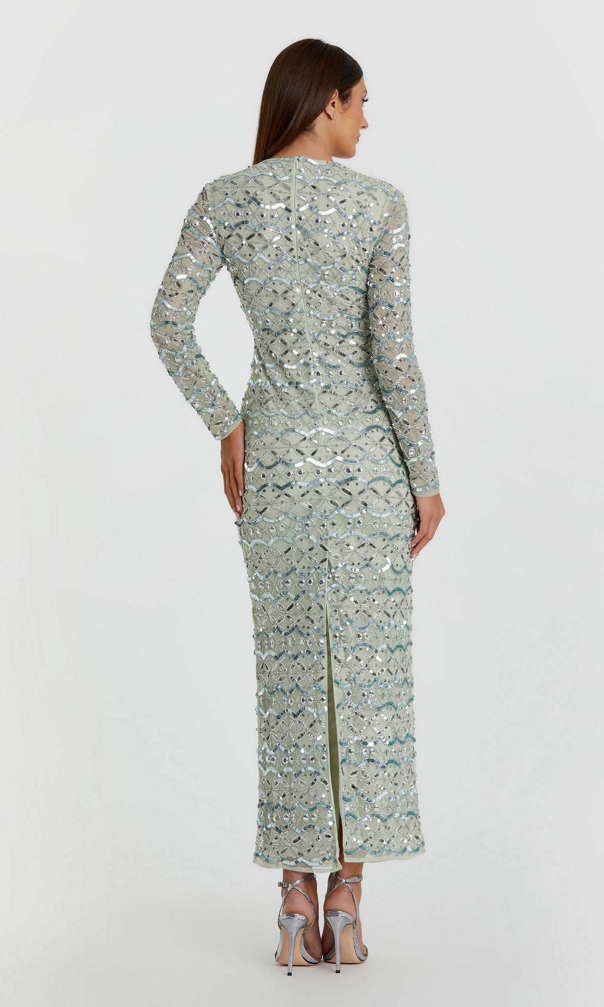 A woman faces away, wearing the Mac Duggal 94194 long formal dress—a long-sleeved, light green sequin gown with a back slit—paired with silver high-heeled sandals against a plain background.