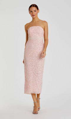 A woman poses in the Tea-Length Party Dress: Mac Duggal 94200—a blush, sequined midi design featuring a strapless neckline, fitted silhouette, and subtle pink pearl-embellished details. She completes the elegant look with silver high-heeled sandals against a plain light background.