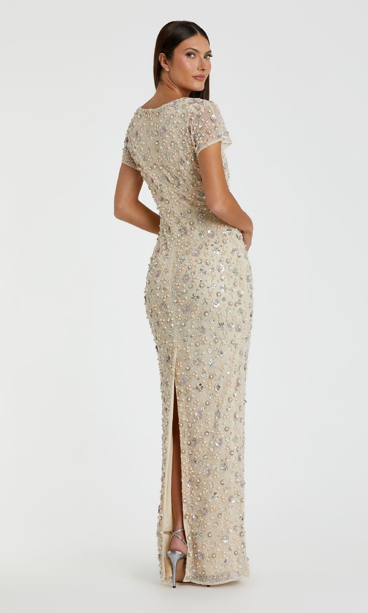 A woman poses with her back turned, looking over her shoulder, wearing the Mac Duggal 94205 Embellished Long Formal Dress in beige—floor-length, fitted, beaded with sequins, short sleeves, and a back slit.