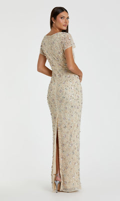 A woman poses with her back turned, looking over her shoulder, wearing the Mac Duggal 94205 Embellished Long Formal Dress in beige—floor-length, fitted, beaded with sequins, short sleeves, and a back slit.