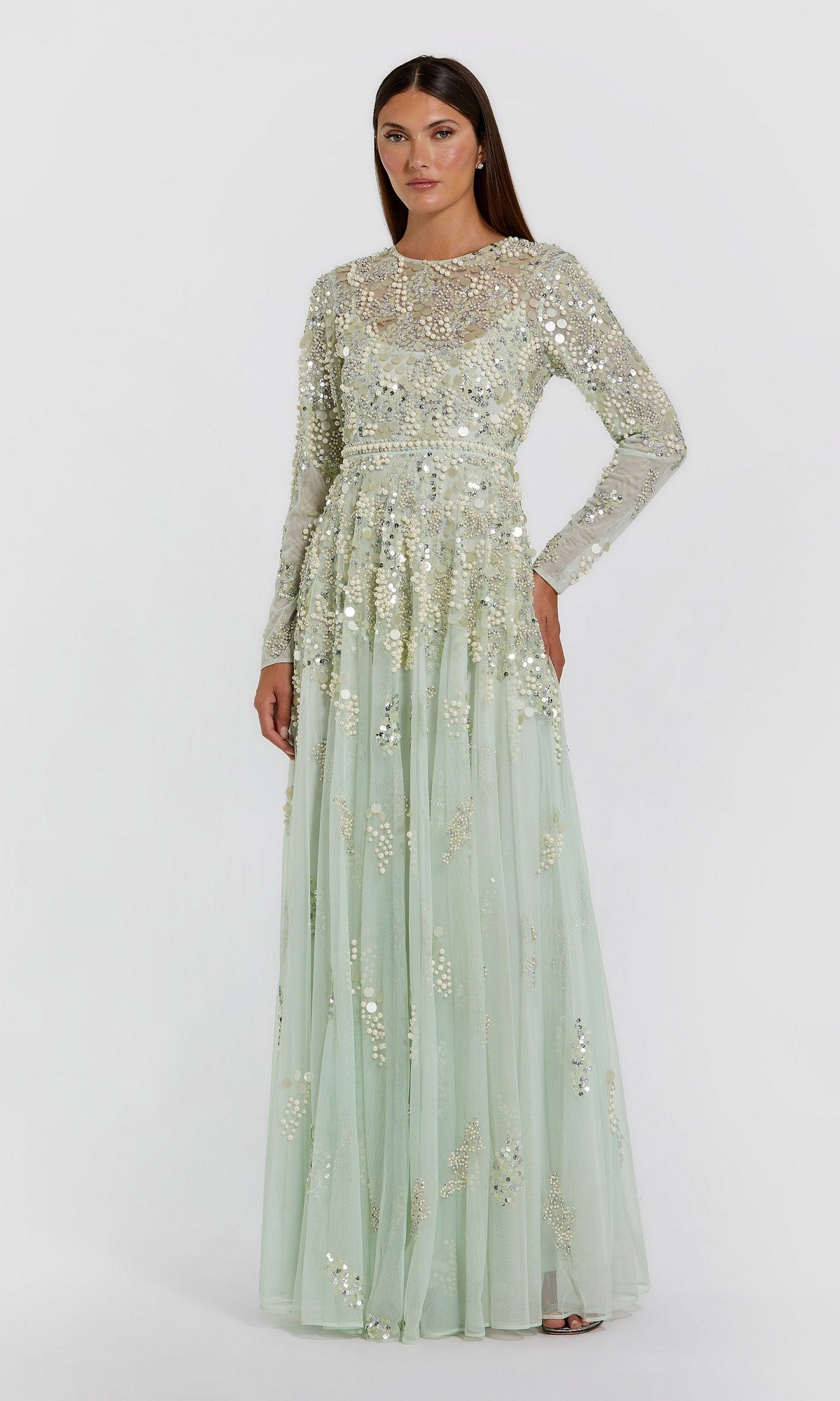 A woman wearing the Mac Duggal 94215 long formal dress in sage, featuring sheer long sleeves and silver sequin embellishments, stands against a plain white background.