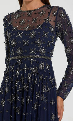 Wearing the Mac Duggal 94216 Long Formal Dress, a woman showcases a navy blue sheer gown adorned with silver beaded stars and cross patterns, featuring long sleeves, a fitted waist, and exquisite beaded embellishments.