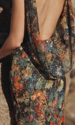 A person wearing the Mac Duggal 94263 long formal dress, featuring sheer floral mesh and a low-cut back, stands against a textured stone surface.