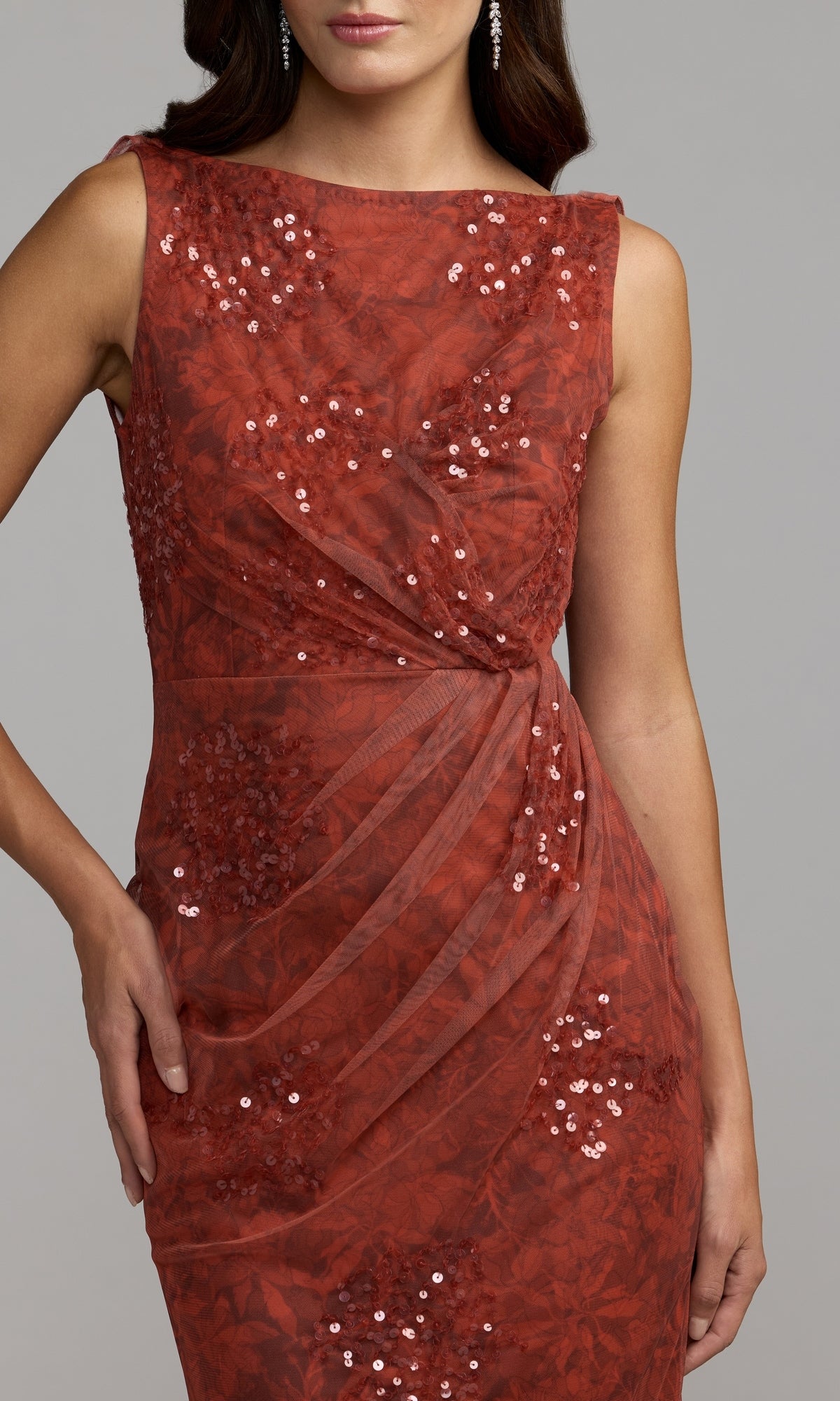 A woman poses with one hand on her hip against a plain background, wearing the Mac Duggal 94263 Cowl-Back Floral Formal Gown—a sleeveless, fitted rust-red sequin dress featuring a subtle floral pattern.