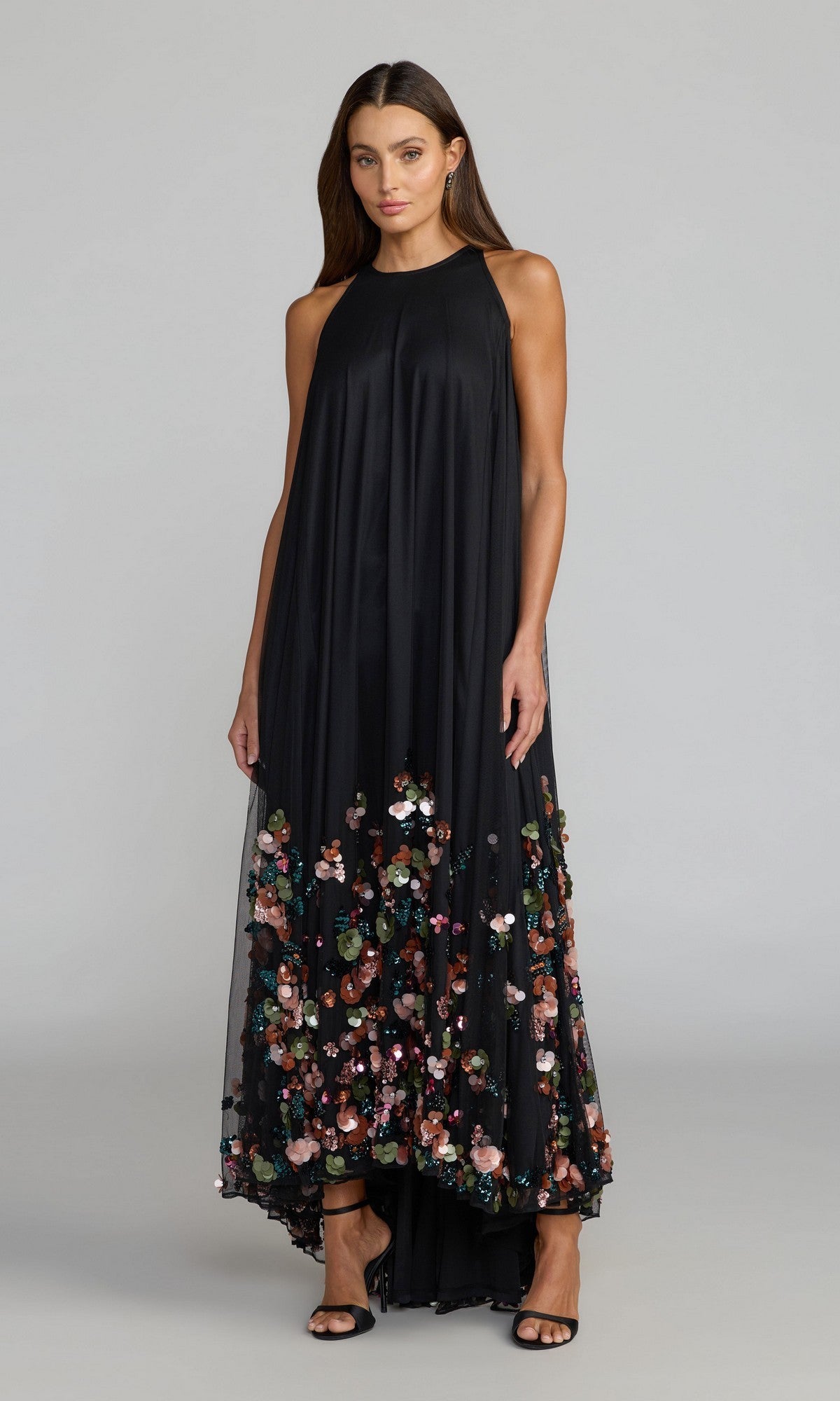 A woman stands against a plain gray background wearing the Mac Duggal 94270 Long Formal Dress in Black Multi—a sleeveless black mesh gown with a high neckline and colorful 3D sequin floral accents near the hem—paired with black heeled sandals.