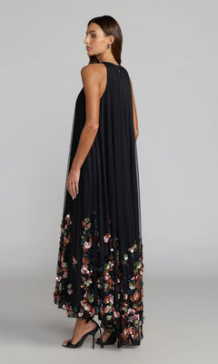 A woman in profile stands against a plain light gray background wearing the Mac Duggal 94270 long formal dress—a sleeveless black mesh gown featuring colorful 3D sequin flower embellishments along the hem.