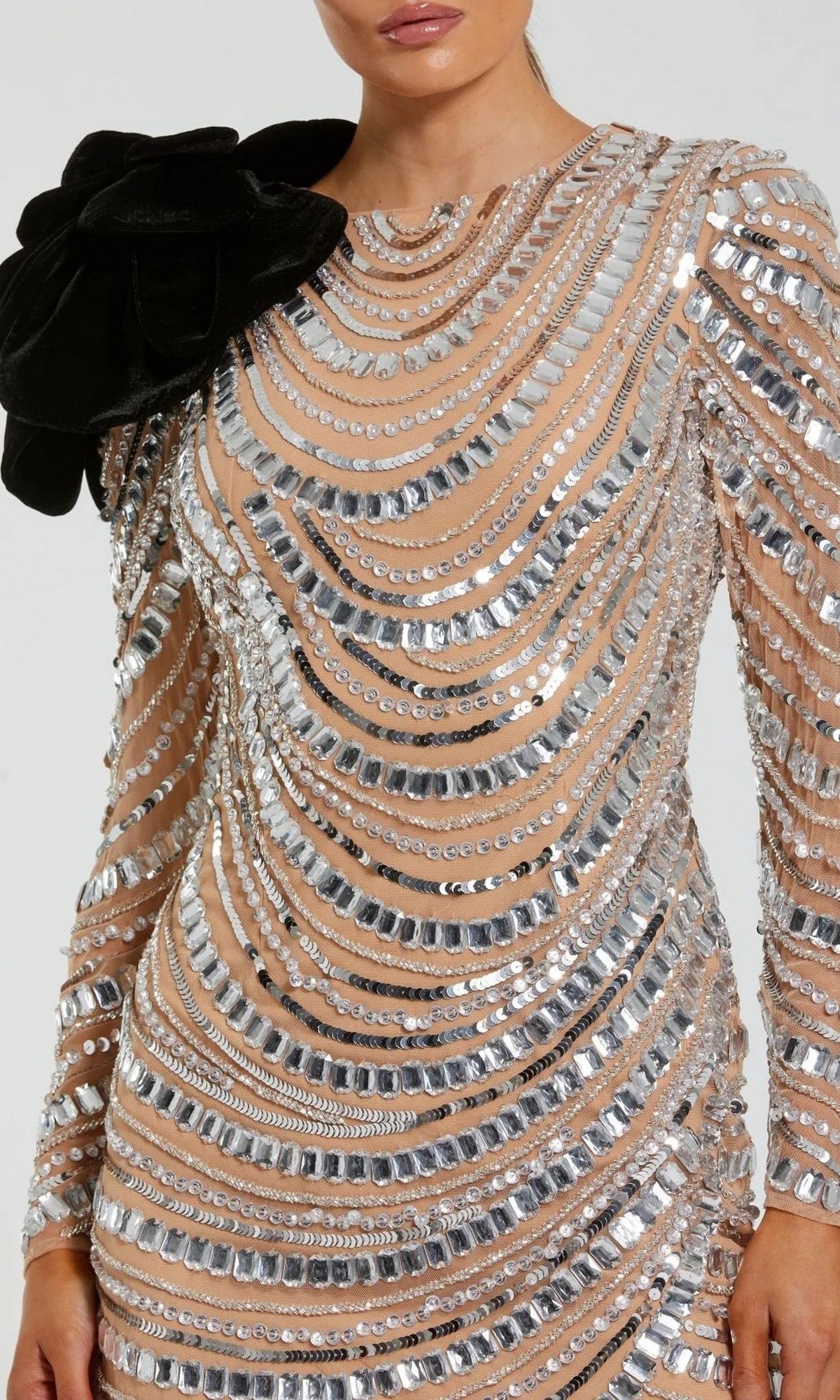 A woman wearing the Mac Duggal 94284 short party dress, featuring nude fabric, long sleeves, silver sequin stripes, and a large black fabric flower on the left shoulder.