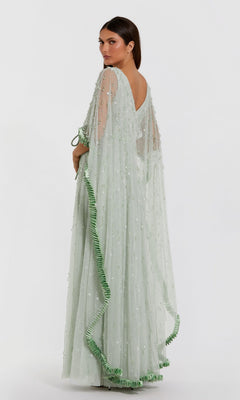 A woman stands against a white background, looking over her shoulder while wearing the Mac Duggal 94294 Long Formal Dress—a light green sheer gown featuring embroidered details and ruffled trim.