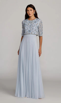 A woman stands against a plain background wearing the Mac Duggal 9430 long formal dress, featuring a powder blue chiffon A-line skirt that falls to the floor, with floral embroidery on the bodice and sheer half sleeves.