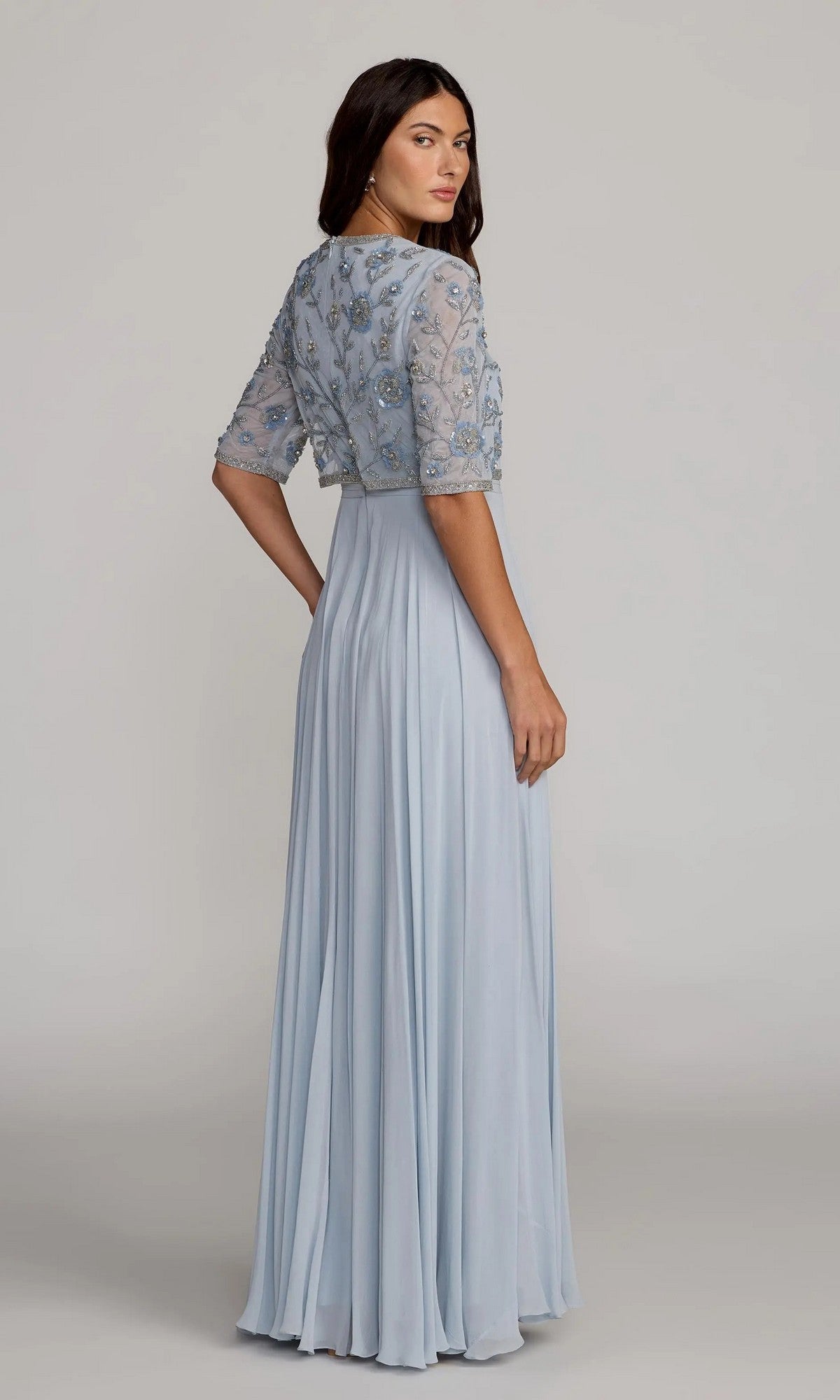 A woman stands against a plain background, looking over her shoulder while wearing the Mac Duggal 9430—a long formal dress featuring a light blue floor-length chiffon A-line skirt and an embroidered sheer top adorned with floral designs.