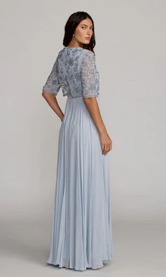 A woman stands against a plain background, looking over her shoulder while wearing the Mac Duggal 9430—a long formal dress featuring a light blue floor-length chiffon A-line skirt and an embroidered sheer top adorned with floral designs.