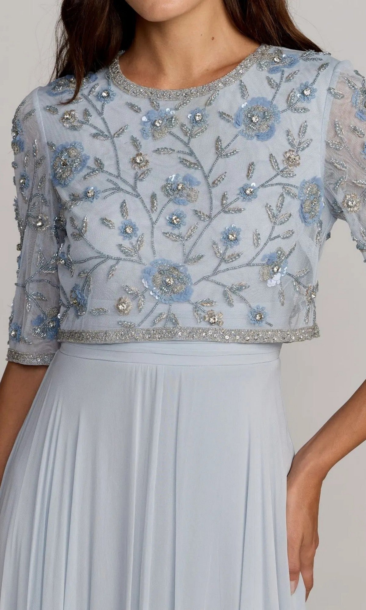 A woman stands with one hand on her hip, wearing the Mac Duggal 9430 long formal dress—a light blue chiffon A-line gown with sheer short sleeves and delicate floral embroidery adorning the bodice.