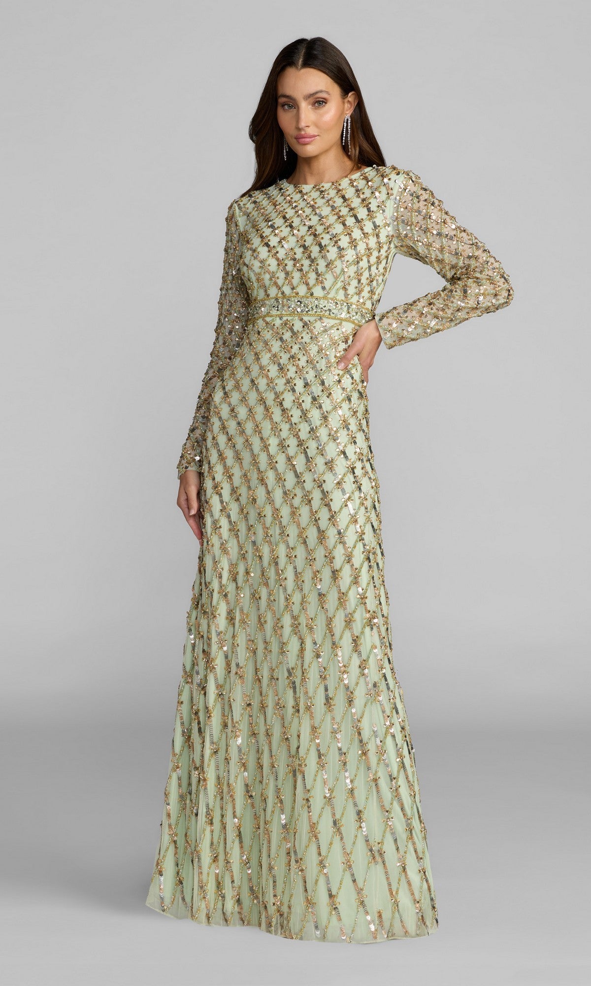 A woman stands against a plain gray background wearing the Mac Duggal 94318 Long Formal Dress in sage, featuring modest long sleeves, a full-length silhouette, and a diamond-patterned overlay adorned with sequin embellishments.