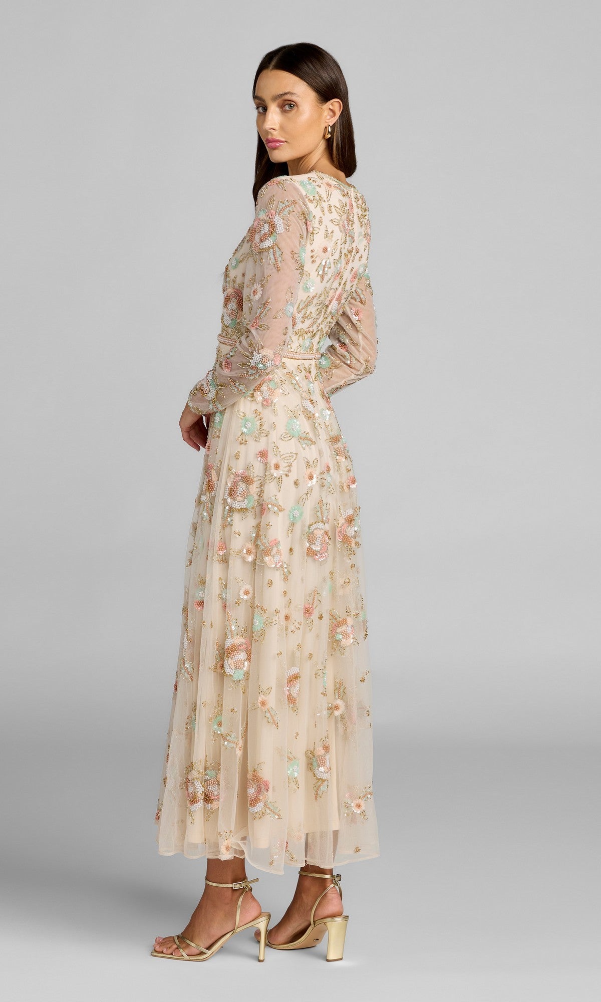 A woman wearing the Mac Duggal 94336 ankle-length floral dress—featuring sheer long sleeves, a sheer cream fabric, and beaded floral embellishments—stands sideways in gold high-heeled sandals against a plain gray background.